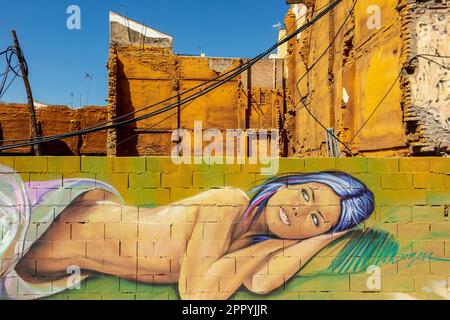 Wandgemälde einer liegenden jungen Frau, gemalt an einer Wand in Malaga Andalusien, Spanien, mit zerstörtem Gebäude im Hintergrund und blauem Himmel darüber. Stockfoto