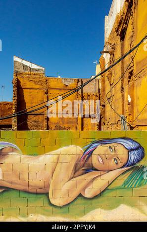 Wandgemälde einer liegenden jungen Frau, gemalt an einer Wand in Malaga Andalusien, Spanien, mit zerstörtem Gebäude im Hintergrund und blauem Himmel darüber. Stockfoto