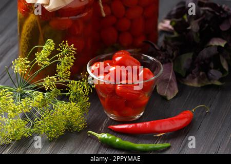 Eingelegte Kirschtomaten in einer Schüssel, Knoblauch, rote und grüne Chili-Paprika, Dill, Basilikum auf einem hölzernen Hintergrund, Gläser mit Tomatenkonserven Stockfoto