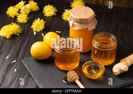 Honig in Gläsern, Dipper, Zitrone, Ingwer auf einer schwarzen Steinplatte auf Holzhintergrund. Der Begriff der Bienenprodukte. Stockfoto