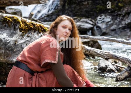 Das rothaarige, langhaarige Modell sitzt am Ufer eines Bergflusses und schaut direkt in die Kamera Stockfoto
