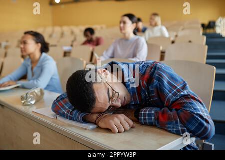 Teenager, der während des Vortrags am Schreibtisch schläft, während andere Schüler im Hintergrund sitzen Stockfoto