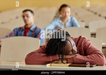 Der afroamerikanische Schuljunge liegt auf dem Schreibtisch und schläft während einer langweiligen Vorlesung mit seinen Klassenkameraden im Hintergrund Stockfoto