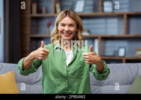 Porträt einer glücklichen älteren Frau in einem grünen Kleid, die zu Hause auf der Couch sitzt und lächelt und Superhände vor der Kamera zeigt. Stockfoto