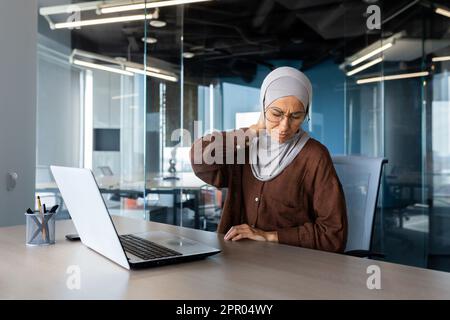 Müde junge arabische Geschäftsfrau in Hijab, die an Projektüberstunden im Büro mit Laptop arbeitet und Nacken hält, Schmerzen grimpert, mit Händen massiert. Stockfoto