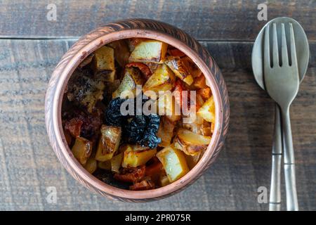 Gedünstete Kartoffeln, Karotten, Zwiebeln, Tomaten und Backpflaumen in einem Tontopf auf Holztisch-Hintergrund, Nahaufnahme, Draufsicht Stockfoto