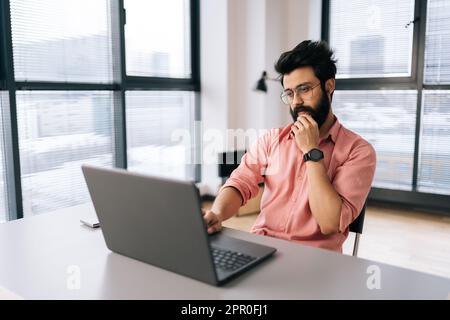 Porträt eines pensiven indischen Freiberuflers, männlich, der am Schreibtisch sitzt und sich mit dem Kopf an die Hand lehnt und auf den Bildschirm schaut. Stockfoto
