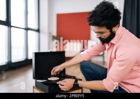 Porträt eines bärtigen indischen Hipsters in Brille, der den Kopf eines alten Plattenspielers justiert, und Vinylscheiben, die auf dem Boden liegen. Musikliebhaber, der zuhört Stockfoto
