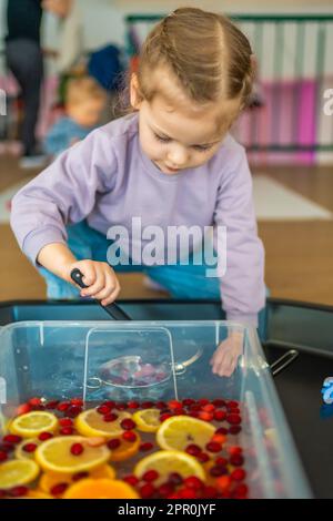 Kleines Mädchen, das in einer handgemachten Obstbar mit Küchenutensilien, Beeren und Zitrusfrüchten spielt. Sensorische Entwicklung und Erfahrungen, Themenaktivitäten mit Stockfoto