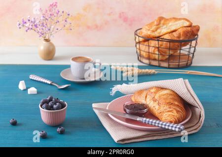 Leckeres Frühstück mit Croissants, frischen Heidelbeeren, Marmelade und einer Tasse Kaffee. Blauer Holztisch, niedriger Blickwinkel, keine Menschen. Stockfoto