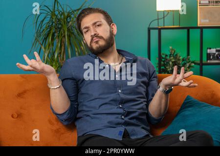 Ich weiß nicht, was ich sagen soll. Verwirrter, gutaussehender mann im libanon, der sich wegen einer zweideutigen Frage schämt, Zweifel, keine Ahnung hat, zu Hause ahnungslos und unsicher ist. Der Arabier sitzt auf dem Sofa im Zimmer Stockfoto