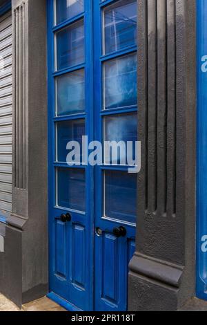 Blaue Doppeltür aus Holz mit Glasfenstern, geschlossenen Fensterläden vor dem Hintergrund grauer Wände Stockfoto