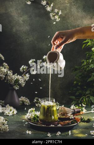 Die Frauenhand gießt hausgemachte Sauerrahm aus einem kleinen Glas in ein Glas mit Matcha-Tee. Dunkler Hintergrund mit Frühlingskirschblüte und traditionellen Gerichten Stockfoto