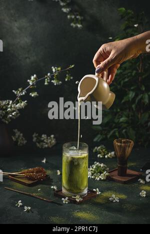 Die Frauenhand gießt hausgemachte Sauerrahm aus einem kleinen Glas in ein Glas mit Matcha-Tee. Dunkler Hintergrund mit Frühlingskirschblüte und traditionellen Gerichten Stockfoto