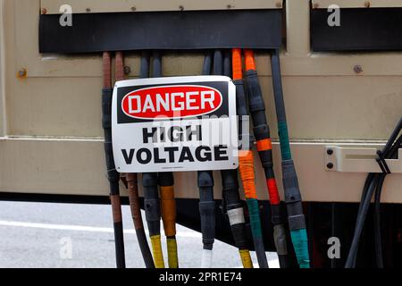 Ein Schild mit der Aufschrift „Gefahr durch Hochspannung“, das auf elektrischen Leiterkabeln angebracht ist, die einen Mietgenerator verlassen, sind die Kabel in amerikanischer brauner orangefarbener gelber Phasenfarbe Stockfoto