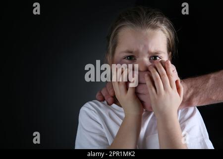 Ein Mann, der den Mund eines verängstigten kleinen Mädchens auf schwarzem Hintergrund verdeckt, Platz für Text. Häusliche Gewalt Stockfoto