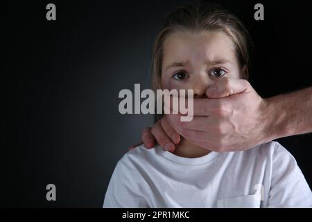 Ein Mann, der den Mund eines verängstigten kleinen Mädchens auf schwarzem Hintergrund verdeckt, Platz für Text. Häusliche Gewalt Stockfoto