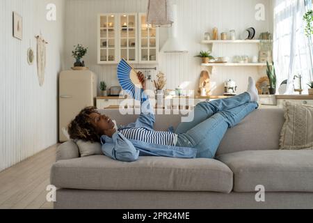 Unglückliche erschöpfte und überhitzte junge afrikanische Frau, die zu Hause auf dem Sofa liegt und mit dem Ventilator winkt, um sich abzukühlen Stockfoto