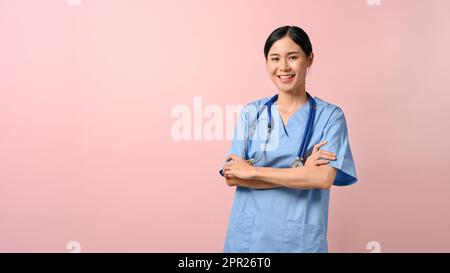Porträt einer jungen, attraktiven Ärztin, die steht und ihre Arme vor einem pinkfarbenen isolierten Hintergrund kreuzt. Stockfoto