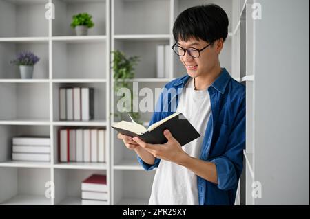 Junger asiatischer Student mit Brille, der im Arbeitszimmer Buch liest, lehnt sich gegen das Bücherregal. Stockfoto