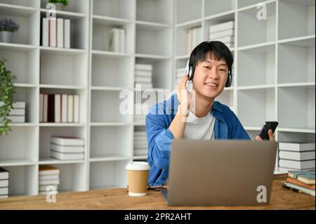 Junger, bezaubernder asiatischer Mann, der Musik mit Kopfhörern hört, während er im Wohnzimmer einen Laptop benutzt. Stockfoto