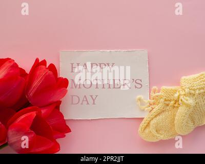 Rote Tulpenblumen und gelbe Schühchen für ein Neugeborenes auf pinkfarbenem Hintergrund mit Happy Mütters Day. Muttertag Grußkarte. Wünsche für Mutter schreiben. K Stockfoto