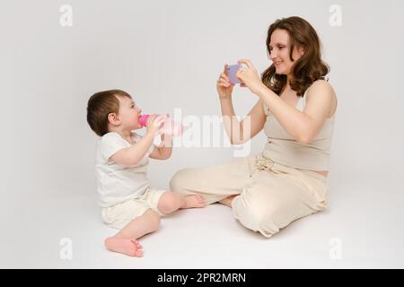 Die Mutter macht ein Foto von einem Baby mit einem Telefon auf weißem Hintergrund. Die Bloggerin der Mutter nimmt ein Video von einem Kind für einen Blog auf. Der Junge ist ungefähr zwei Yen Stockfoto