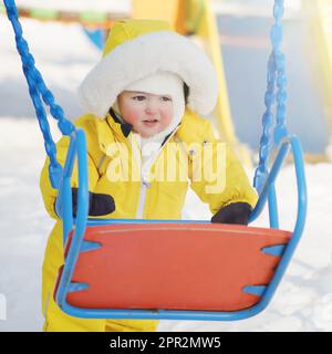 Ein glückliches Kleinkind klettert auf dem Winterspielplatz selbst auf die Schaukel. Ein Junge im gelben Schneeanzug, der im Kindergarten schwingt. Ein Kind im Alter von acht Jahren Stockfoto