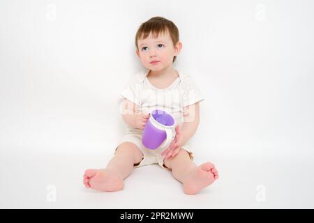 Fröhliches Baby trinkt Wasser aus einer Tasse auf weißem Studiohintergrund. Ruhendes Kind mit Tassensaft in den Händen. Kind etwa zwei Jahre alt (ein Jahr neun Monate) Stockfoto