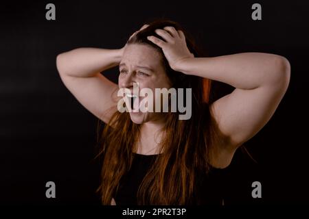 Junge verärgerte Frau mit langen Haaren. Schrei und schreie laut mit offenem Mund. Weibliches Porträt isoliert auf Schwarz, Seitenansicht Stockfoto