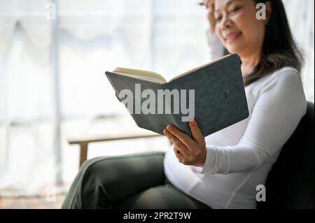 Eine wunderschöne asiatische Frau mittleren Alters liest ein Buch, während sie zu Hause auf dem Sofa sitzt Stockfoto