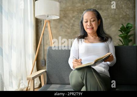 Eine wunderschöne asiatische Frau mittleren Alters liest ein Buch, während sie zu Hause auf dem Sofa sitzt. Lifestyle-Konzept Stockfoto