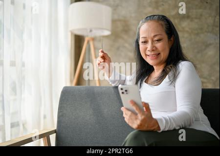 Glückliche ältere Frau, die mit ihrem Smartphone online einkauft und dabei auf dem Sofa sitzt. Stockfoto