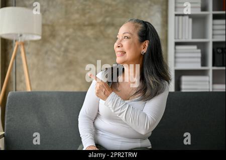 Attraktive und lächelnde asiatische Erwachsene Frau sitzt auf dem Sofa in ihrem Wohnzimmer, schaut in die Ferne und zeigt mit dem Finger zur Seite. Stockfoto