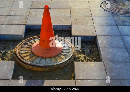 Ein leuchtend orangefarbener Kegel steht auf dem Abfluss. Markierung gefährlicher Abschnitte auf der Straße, Zutritt für Personen ist verboten. Der Kegel steht auf dem brok Stockfoto