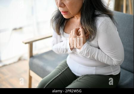 Nahaufnahme einer unwohltuenden und ungesunden asiatischen, reifen Frau, die in ihrem Wohnzimmer auf dem Sofa sitzt und an Herzinfarkt oder Brustschmerzen leidet. Stockfoto