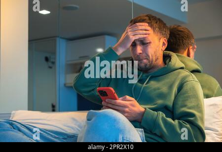 Ein Mann in Schwierigkeiten reagiert emotional auf schlechte Nachrichten, die er am Telefon gelesen hat oder die sich wegen des Anrufs gestritten haben. Deprimierter Kerl zu Hause mit Smartphone auf dem Bett. Stockfoto