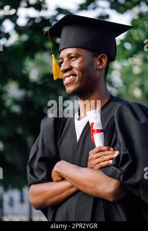 Portrait Side View eines glücklichen afro-amerikanischen Absolventen von der Universität stehen im Freien mit gekreuzten Armen und haben ein Hochschuldiplom, selektiv Stockfoto