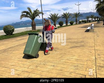 Ein männlicher Putzmann in Uniform, ein Bademantel, der einen Spaten auf seiner Schulter hält und einen Mülleimer auf Rädern in einem tropischen, warmen Paradies-Resort herumschleppt Stockfoto