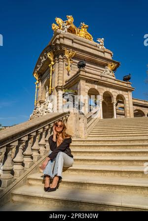 Junge, gebräunte, schöne Frau mit geschlossenen Augen in einem schwarzen Oberteil und Jeans mit Sonnenbrille und langen Haaren, die stilvoll und elegant in der Sonne sitzt Stockfoto