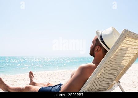Junger Mann mit weißem Hut, der sich auf einem Liegestuhl am wunderschönen Sandstrand entspannen kann. Sommerferienkonzept. Mann entspannt am Strand, Meerblick, Dominikanische Republik Stockfoto