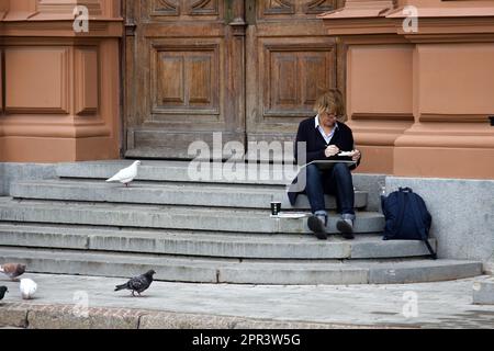 Lettland, Riga - 4. September 2017: Der Künstler sitzt auf der Treppe zum Eingang des Gebäudes. Weiße Tauben sind hier Stockfoto