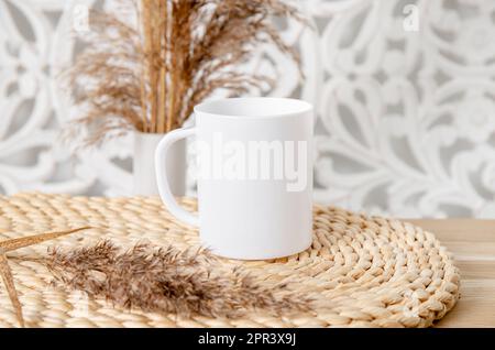 Ein weißes, leeres Tassenmodell im Wohnzimmer. Eine Tasse auf einem Holztisch mit trockener Schilfpflanzendekoration. Viel leerer Kopierspeicher. Stockfoto