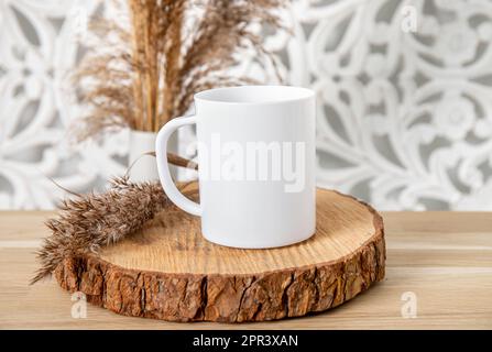 Ein weißes, leeres Tassenmodell im Wohnzimmer. Eine Tasse auf einem Holztisch mit trockener Schilfpflanzendekoration. Viel leerer Kopierspeicher. Stockfoto