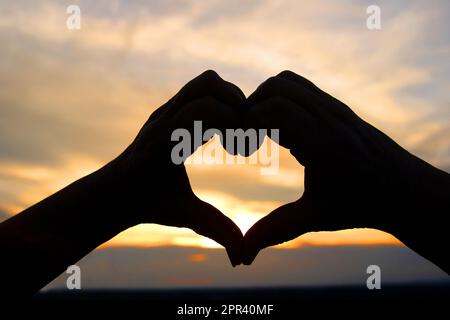 Hände bilden ein Herz vor einem romantischen Sonnenuntergang Stockfoto