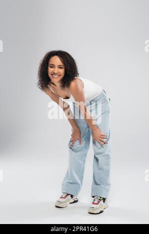 Volle Länge einer sorgenfreien afroamerikanischen Frau in blauen Jeans, die vor der Kamera lächelt, auf grauem Hintergrund, Stockbild Stockfoto