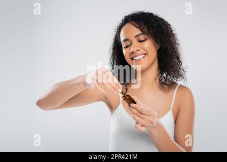 Brünette afroamerikanische Frau in weißem Tank, Öffnung kosmetisches Serum isoliert auf grauem Stock Bild Stockfoto