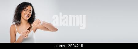Brünette afroamerikanische Frau in einem kosmetischen Serum, isoliert auf grauem Banner, Stockbild Stockfoto
