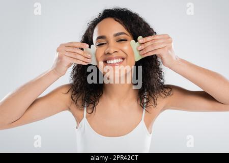 Brünette afroamerikanische Frau, die Gesichtsschaber benutzt, isoliert auf grauem Stockbild Stockfoto