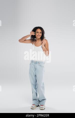 Lange, lächelnde afroamerikanische Frau in Tanktop-Top und Jeans posieren mit Gesichtswäscher und Jade-Rolle auf grauem Hintergrund, Stockbild Stockfoto